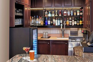 Zona de bar con diversas botellas de licor, un refrigerador, una grifería y una copa de martini con una rodaja de limón.