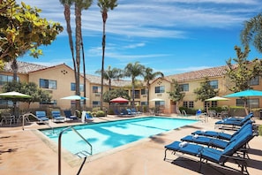 A pool area with lounge chairs and umbrellas, surrounded by palm trees and a building with a tiled roof. - Spacious Open-Plan King Premier | Poolside Access | 0.1 Mi to Magic Mountain (Valencia)