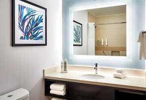 Un baño con lavamanos, espejo, una obra de arte enmarcada y artículos de aseo.
