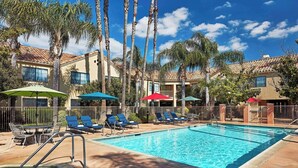 Una piscina con sillas de descanso y sombrillas, rodeada de palmeras y un edificio con techo de tejas.