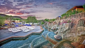 A pool area with lounge chairs and umbrellas, surrounded by rocky terrain and a sunset sky.