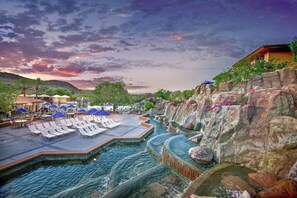 A pool area with lounge chairs and umbrellas, surrounded by rocky terrain and a sunset sky.