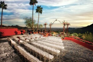 Un espacio para bodas al aire libre con filas de sillas blancas, piso de piedra y un fondo de montañas y palmeras.