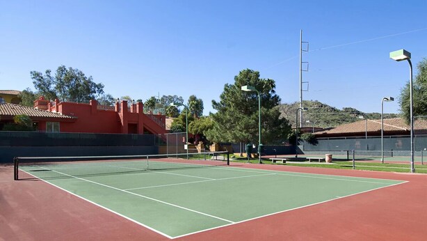 An outdoor tennis court with a net, surrounded by a fence, and a building with a red roof in the background.