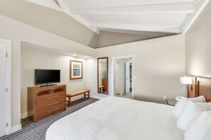 Een hotelkamer met een groot bed, een televisie op een houten kast, een bankje, een spiegel en een deur naar een kast.