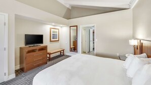 Ein Hotelzimmer mit einem großen Bett, einem Fernseher auf einer Holzkommode, einer Bank, einem Spiegel und einer Tür zu einem Kleiderschrank.