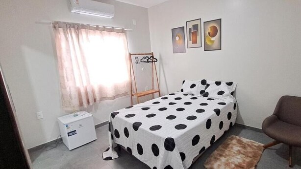 Quarto com cama, cadeira, mesa de cabeceira, aquecedor e janela com cortinas.