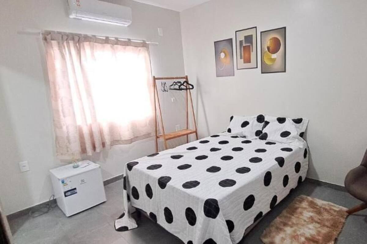 Un dormitorio con cama, una silla, una mesita de noche, un calentador y una ventana con cortinas.