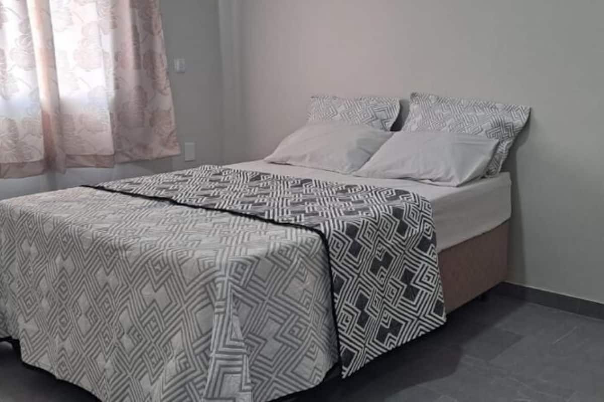 Una cama con ropa de cama estampada, una mesita de noche y una ventana con cortinas.