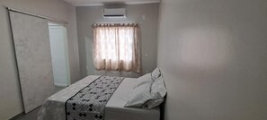 A bedroom with a bed, a window with curtains, an air conditioning unit, and a door. - HOSPEDARIA LENÇÓIS  (Barreirinhas)
