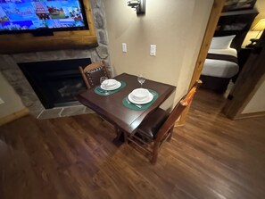 A dining area with a wooden table set for two, a stone fireplace, and a television mounted on the wall.