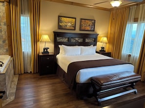 Hotelkamer met een groot bed, twee bedlampen, een hoofdeinde met kunstwerk, een bankje en een stenen muur.