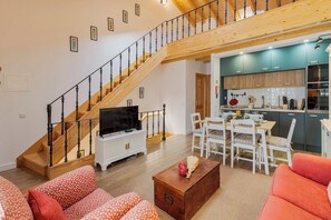Ein geräumiger Wohnbereich mit Holzboden und einer Wendeltreppe, einem Esstisch mit Stühlen, einem Fernseher auf einem Schrank und einer Küche im Hintergrund.