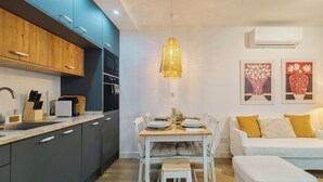 A modern kitchen with dark cabinets, a dining table set for four, and a white sofa with yellow cushions. - Casa do Bocage, By Home Match (Setúbal)
