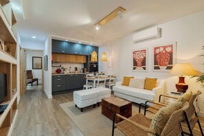 A modern living room with a sofa, ottomans, and wicker chairs. There is a dining area with a table and chairs, and a kitchen with dark cabinets and a hanging light fixture. - Casa do Bocage, By Home Match (Setúbal)