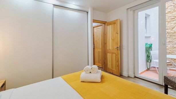 A bedroom with a bed, a yellow rug, a wooden door, and a view of the outside. - Casa do Bocage, By Home Match (Setúbal)