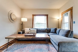 Ein Wohnzimmer mit Ecksofa, Couchtisch aus Holz, gemustertem Teppich, Wandspiegel und Stehlampe.