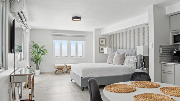 Ein modernes Hotelzimmer mit Bett, Nachttischen, einem Schreibtisch, einem Sessel, einem Esstisch mit Platzsets, einer Pflanze und Blick auf die Stadt.