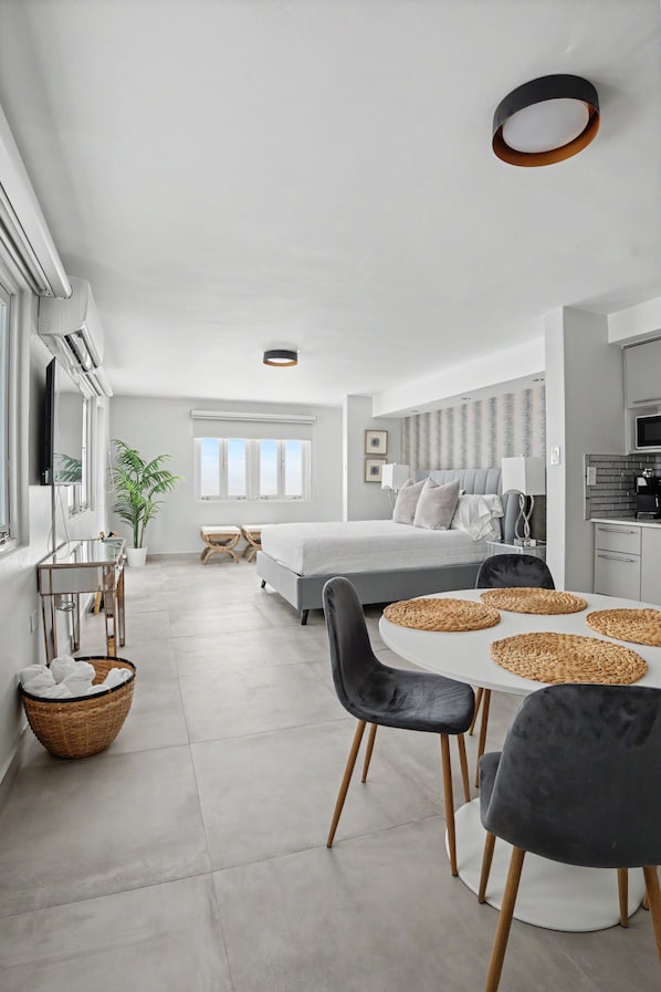 A modern hotel room with a bed, bedside tables, a desk, a chair, a dining table with placemats, a plant, and a view of the city. - Venus Oasis - Pool, Game Room & Full Solar Backup - Steps to the Beach (Carolina)