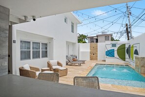 A modern outdoor pool area with wicker furniture, a pool, and a white building with large windows. - Venus Oasis - Pool, Game Room & Full Solar Backup - Steps to the Beach (Carolina)