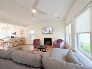 A spacious living area with a fireplace, two armchairs, a sofa, a kitchen with white cabinets, and a ceiling fan. - The Happy Nest - Sleeps 5 Near Kennebunk Beaches (Arundel)
