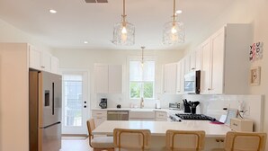A modern kitchen with white cabinetry, a stainless steel refrigerator, a central island with bar stools, pendant lighting, and a view of the outdoors.