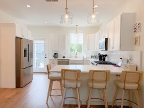 A modern kitchen with white cabinetry, a stainless steel refrigerator, a central island with bar stools, pendant lighting, and a view of the outdoors. - The Maine Happy-Nest / includes Kennebunk Beach Parking (Arundel)