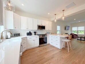 A modern kitchen with white cabinetry, a built-in oven, a microwave, and a gas stove. There are pendant lights, a dining area with a table and chairs, and a large window providing natural light. - The Happy Nest - Sleeps 5 Near Kennebunk Beaches (Arundel)