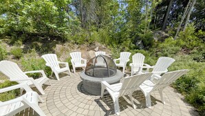 A circular seating area with a fire pit in the center, surrounded by white plastic chairs, set on a paved patio with a natural backdrop.