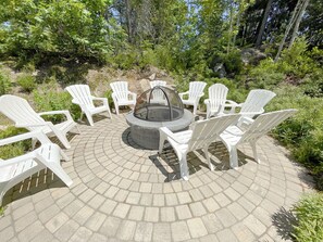 A circular seating area with a fire pit in the center, surrounded by white plastic chairs, set on a paved patio with a natural backdrop. - The Maine Happy-Nest / includes Kennebunk Beach Parking (Arundel)