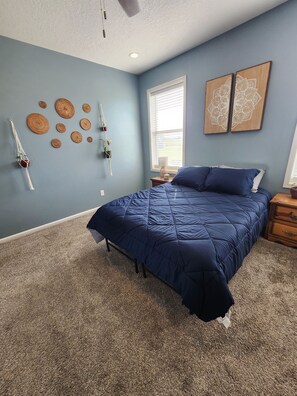 A bedroom with a bed, a window with blinds, a wall-mounted macrame swing, and two wooden wall art pieces. - Group Stays, Sleep 8-10, Near KC, Pet Friendly, 3 acres (Odessa)