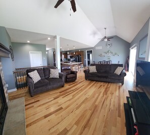 A spacious living room with hardwood floors, two dark sofas with patterned cushions, a ceiling fan, and a kitchen in the background. - Group Stays, Sleep 8-10, Near KC, Pet Friendly, 3 acres (Odessa)