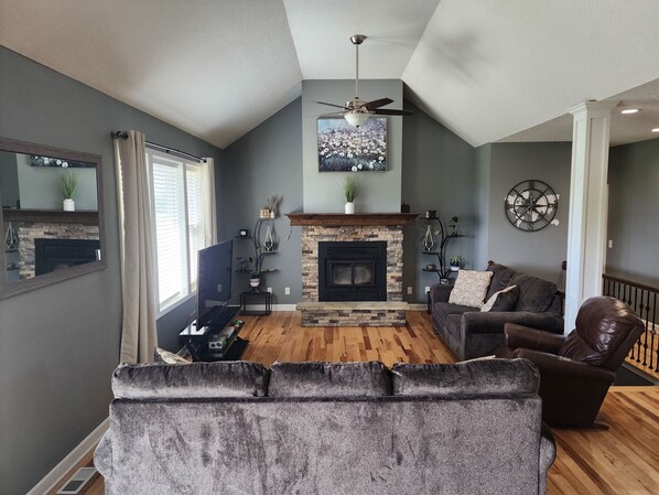 A living room with a fireplace, a ceiling fan, a mounted artwork, and a large mirror.