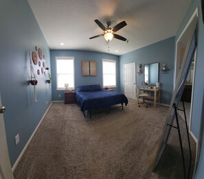 A bedroom with a bed, ceiling fan, two windows, a desk with a chair, and a door.