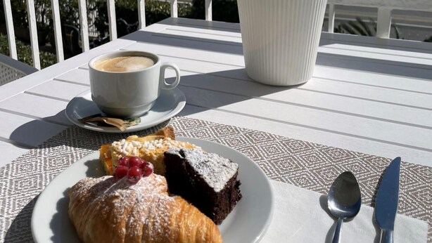 Ein Tisch mit einem Croissant, einer Kuchenstück, einer Tasse Kaffee und Besteck auf einer Terrasse mit Meerblick und einem Hotelschild.