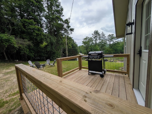 A wooden deck with a grill, chairs, and a view of a wooded area.