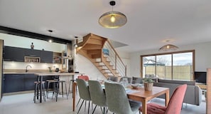 A modern kitchen with a central island, a dining area with a wooden table and chairs, and a staircase leading to an upper level. - La Petite Casa de Silz (ARZAL)