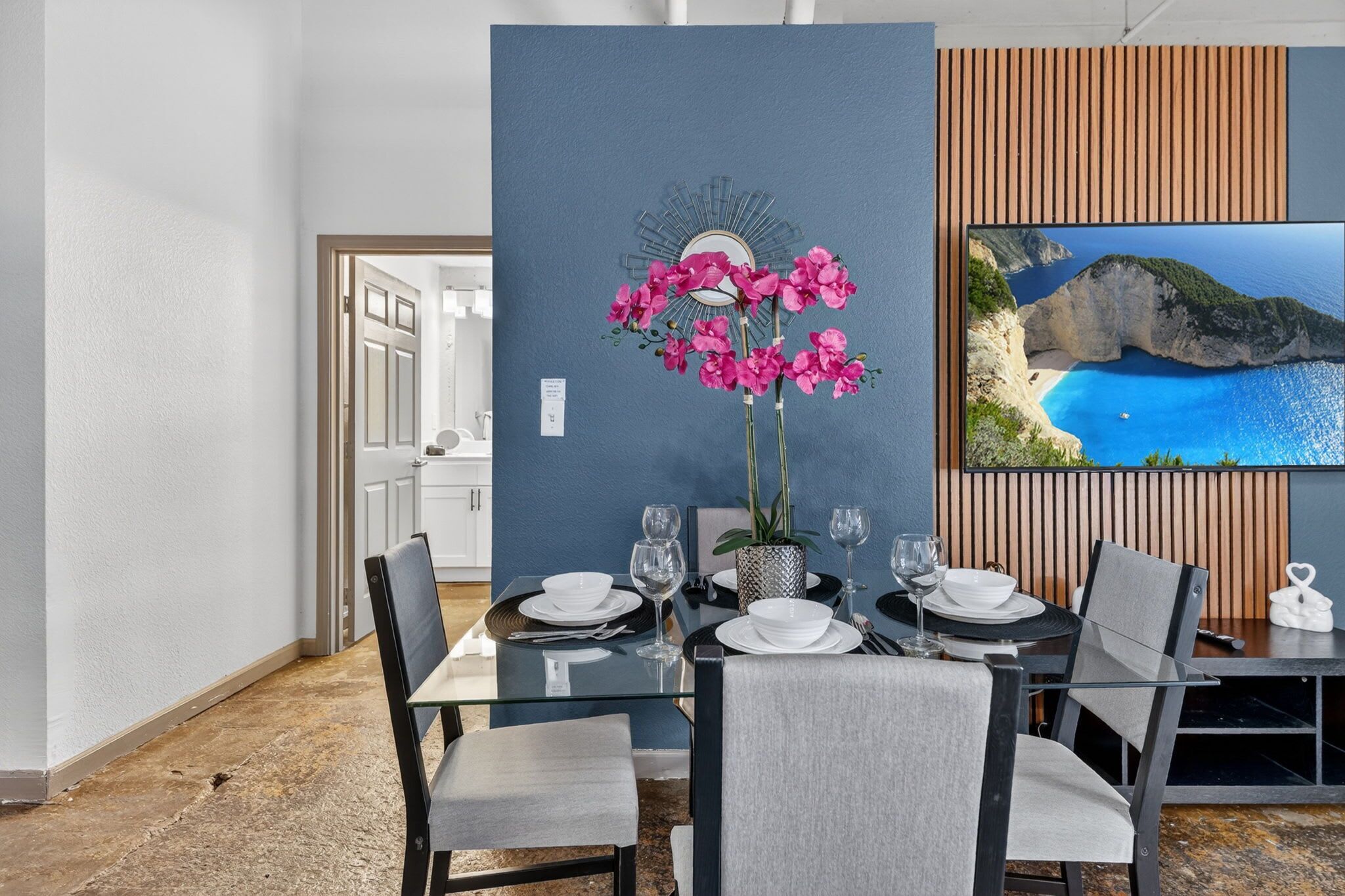A dining area with a table set for four, a vase with pink flowers, and a large framed picture of a coastal landscape.