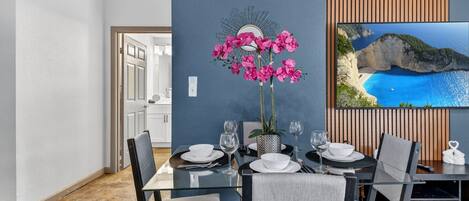 A dining area with a table set for four, a vase with pink flowers, and a large framed picture of a coastal landscape.
