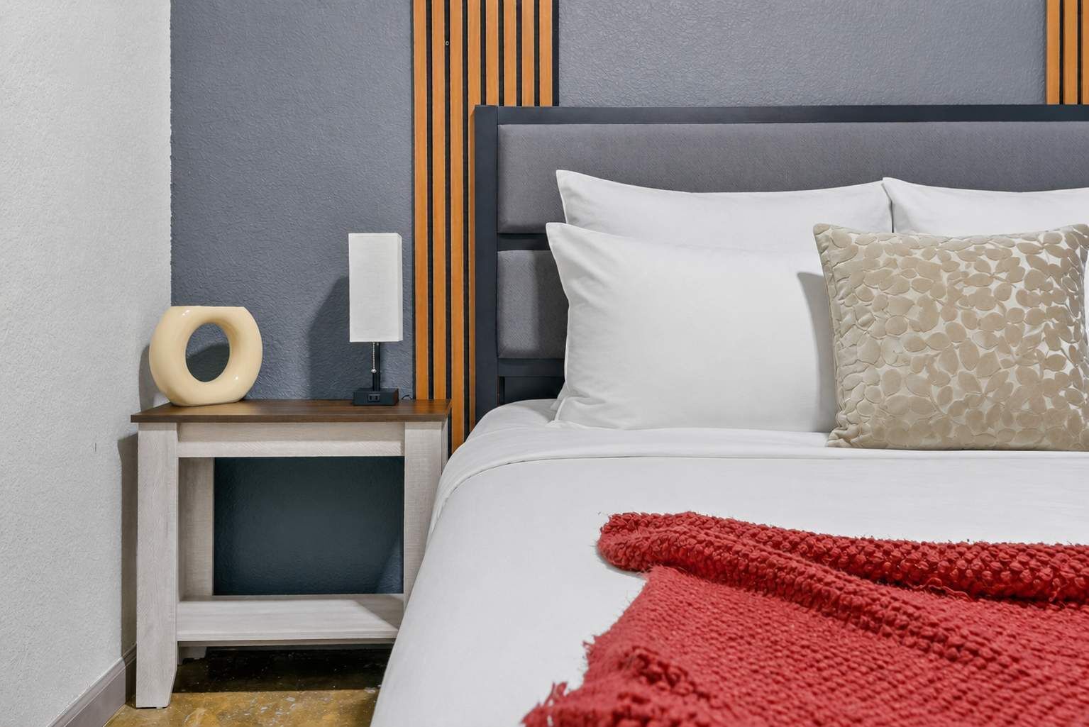 A neatly made bed with white and patterned pillows, a red throw blanket, and a bedside table with a lamp and a decorative object.