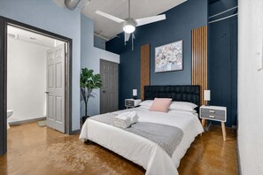 A bedroom with a bed, bedside tables, a ceiling fan, a potted plant, and a painting on the wall. - 2BR Riverfront District Loft | Near Power & Light (Kansas City)