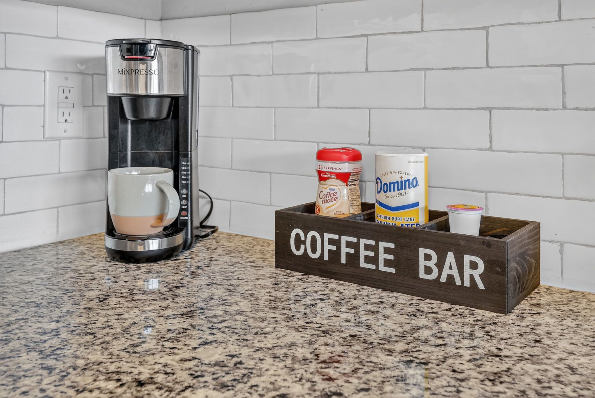 A coffee maker, a coffee mug, a coffee jar, a Domino coffee cup, and a wooden coffee bar sign on a kitchen counter.