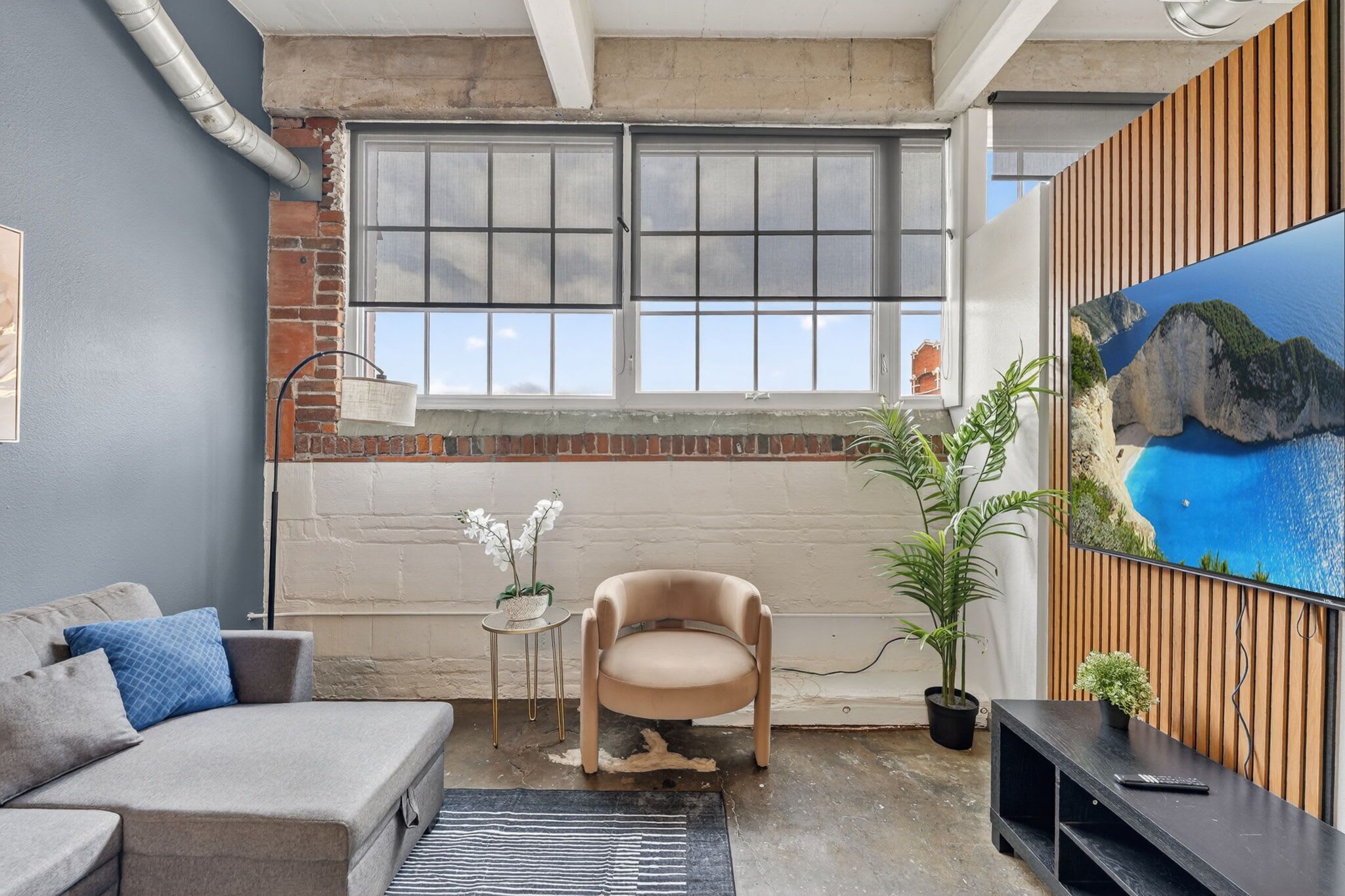 A room with a brick wall, a window, a sofa, a chair, a small table with plants, and a TV on a wooden wall.