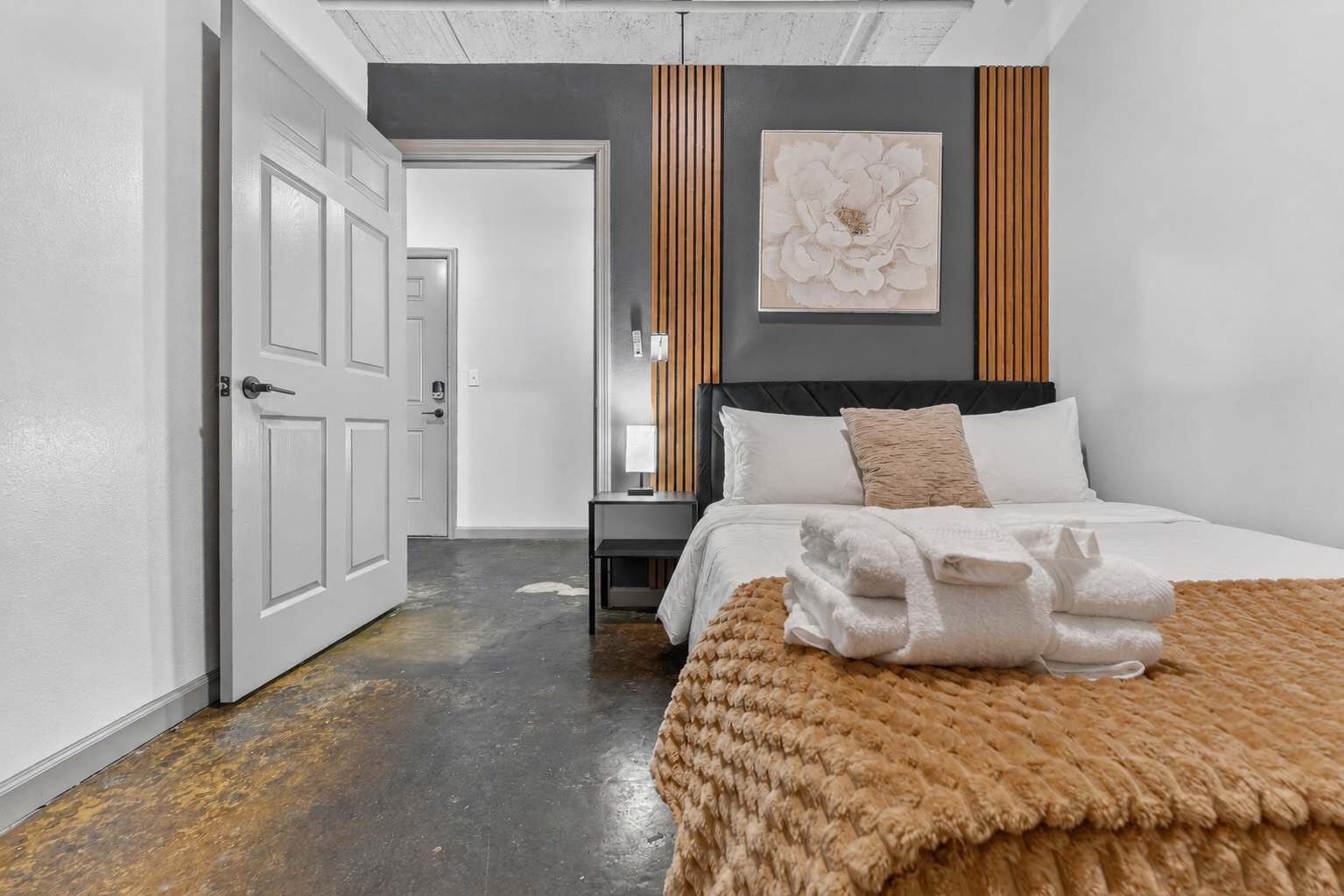 Una habitación de hotel moderna con cama, mesita de noche, una obra de arte colgada en la pared y una puerta que da a otra estancia.