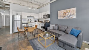 A modern kitchen with stainless steel appliances, a dining area with a glass coffee table, and a grey sofa in a living room.