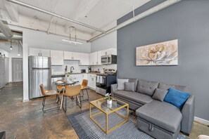 Een moderne keuken met roestvrijstalen apparatuur, een eethoek met een glazen salontafel, en een grijze bank in een woonkamer.