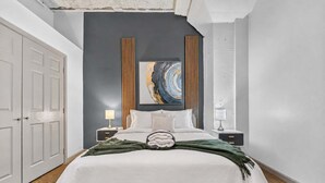 A bedroom with a bed, two bedside tables, a nightstand, a wall-mounted painting, and a door.