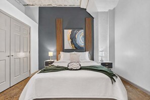 A bedroom with a bed, two bedside tables, a nightstand, a wall-mounted painting, and a door. - River Market Loft Stay - 2BR Stay Near Downtown KC (Kansas City)