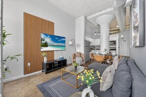 A modern living room with a flat-screen TV, a sofa, a coffee table, and a dining area in the background. - Downtown KC 2BR Loft with Parking & Fast WiFi (Kansas City)