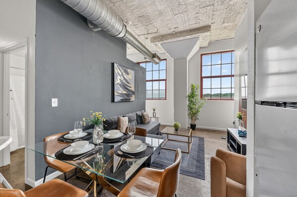 A modern living room with a glass dining table, leather sofas, a large abstract painting, and a view of the outdoors. - Downtown KC 2BR Loft with Parking & Fast WiFi (Kansas City)